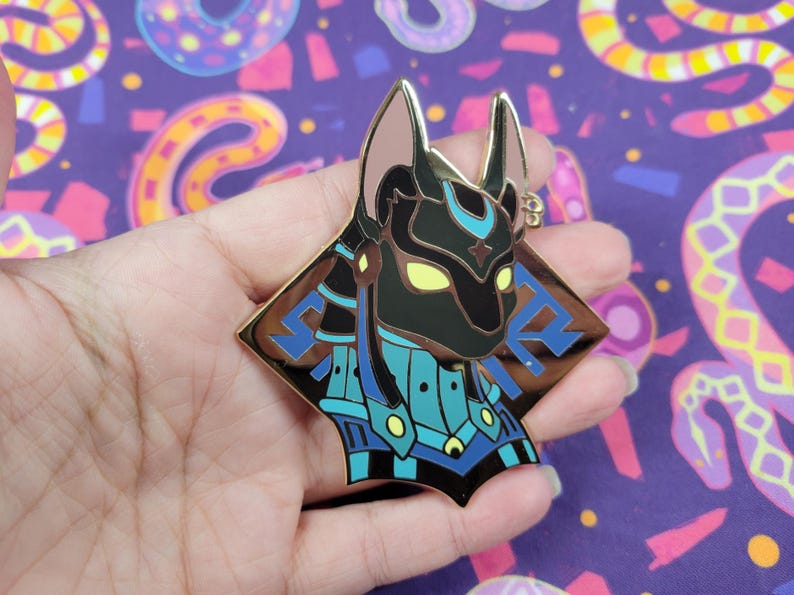 May include: A gold-colored Anubis pin with black, blue, and yellow accents. The pin features a detailed depiction of the Egyptian god Anubis, with a stylized design and a diamond-shaped border. The pin is likely made of metal and has a shiny finish.