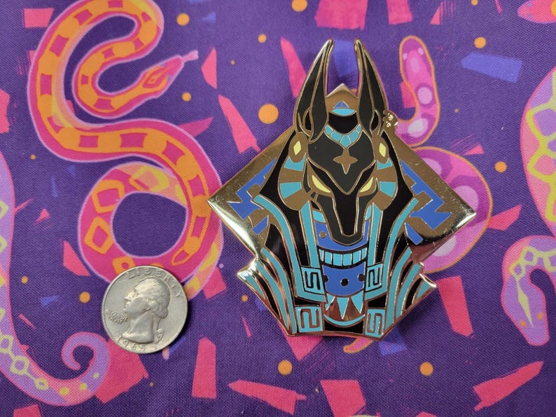 May include: A gold-colored Anubis pin with black, blue, and brown enamel detailing. The pin is diamond-shaped and features the head of the Egyptian god Anubis. The background is a purple fabric with a colorful snake pattern and a quarter coin.