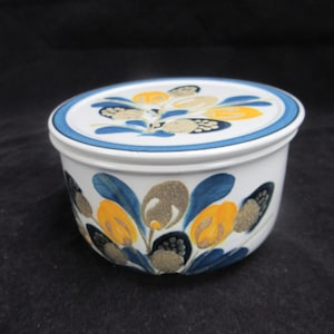 Può includere: Un contenitore in ceramica bianca con coperchio, con un motivo floreale giallo, blu e marrone. Il coperchio ha un bordo blu. Il contenitore rotondo è probabilmente una burriera o un articolo da cucina simile.