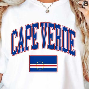 May include: A white t-shirt featuring the text CAPE VERDE in a bold blue font with a red outline. Below the text is a rectangular graphic of the Cape Verde national flag, which consists of blue, white, and red horizontal stripes with yellow stars.