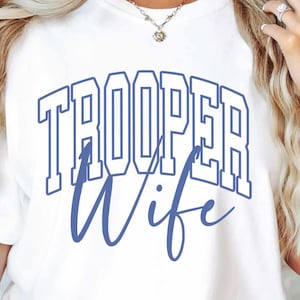 May include: White t-shirt with the words "TROOPER Wife" in blue lettering. The word "TROOPER" is in a block letter style, and "Wife" is in a cursive font. The shirt is short-sleeved.