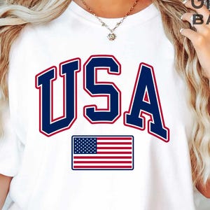 May include: A white short-sleeved t-shirt featuring the text USA in large navy blue letters with a red outline. Below the text is a graphic of the American flag with red and white stripes and a blue rectangle with white stars. The shirt has a crew neckline.