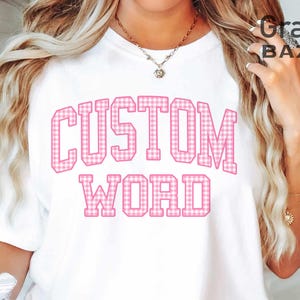 May include: White t-shirt with the words "CUSTOM WORD" in pink, gingham-patterned letters. The shirt has a crew neck and short sleeves. The text is in a bold, collegiate-style font. The image also includes the text "Graphix BAZAAR".