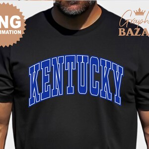 May include: Black t-shirt with the word "KENTUCKY" in blue, outlined in white, in an arched design across the chest. The shirt also has a "PNG SUBMLIMATION" logo in the upper left corner and a "Graphix BAZAAR" logo in the upper right corner.