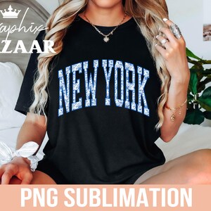 Puede incluir: Camiseta negra con un gr&aacute;fico de "NEW YORK" en purpurina azul y blanca. La camiseta est&aacute; siendo modelada por una persona con cabello largo y rubio.