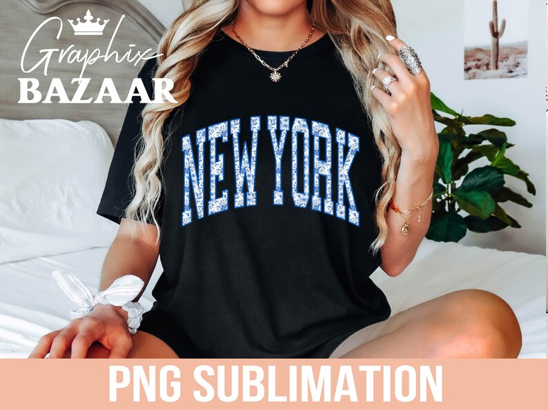 Puede incluir: Camiseta negra con un gr&aacute;fico de "NEW YORK" en purpurina azul y blanca. La camiseta est&aacute; siendo modelada por una persona con cabello largo y rubio.