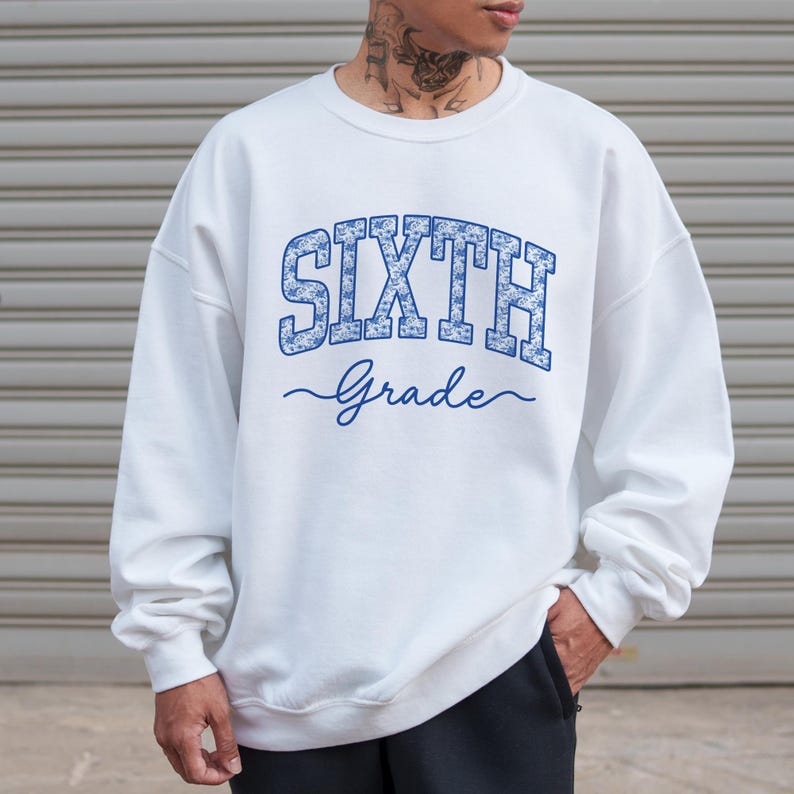 May include: A white crewneck sweatshirt featuring the text "SIXTH" in a blue floral pattern block font, with the word "Grade" written below in a blue cursive script. The garment has long sleeves and a ribbed collar, cuffs, and hem.
