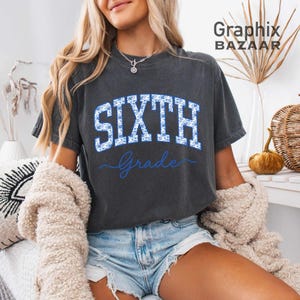 May include: A charcoal gray short-sleeve t-shirt featuring the word SIXTH in large, block-style letters filled with a blue and white floral pattern, with the word Grade written in blue cursive script underneath the block text.