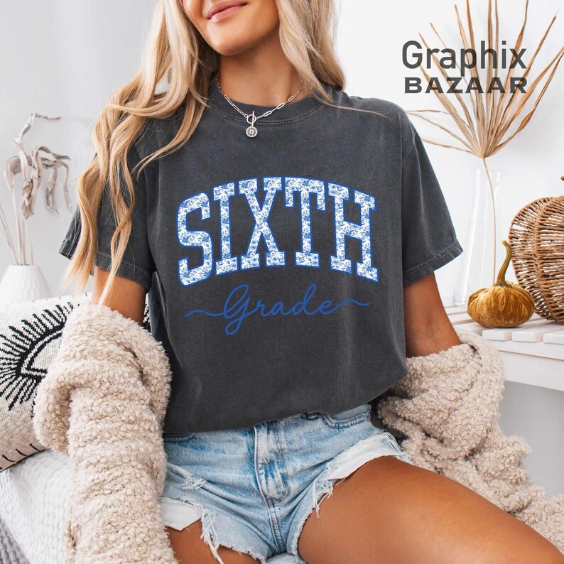May include: A charcoal gray short-sleeve t-shirt featuring the word SIXTH in large, block-style letters filled with a blue and white floral pattern, with the word Grade written in blue cursive script underneath the block text.