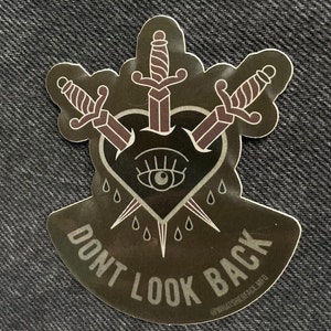 Pode incluir: Um autocolante com um fundo escuro com um coração perfurado por três espadas. O coração tem um olho no centro e lágrimas à sua volta. As palavras "DON'T LOOK BACK" estão impressas na parte inferior.