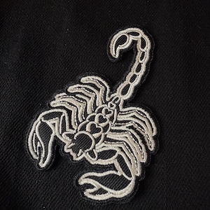 May include: A white embroidered scorpion patch on a black fabric background.
