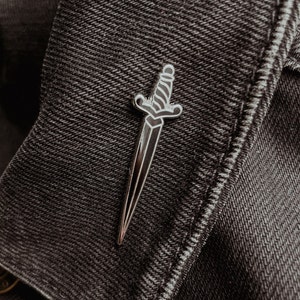 May include: A silver-colored dagger-shaped pin with a black handle and blade, pinned to a dark denim jacket. The pin has a detailed design, including a crossguard and a textured handle. The overall aesthetic is gothic and edgy.