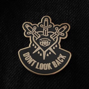 May include: Black and gold enamel pin with a heart pierced by three daggers. The heart has an eye in the center and the text "Don't Look Back" is written on a banner below the heart.