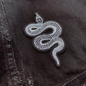 May include: A white snake embroidered patch on a black denim background. The snake is curled up with its head raised.