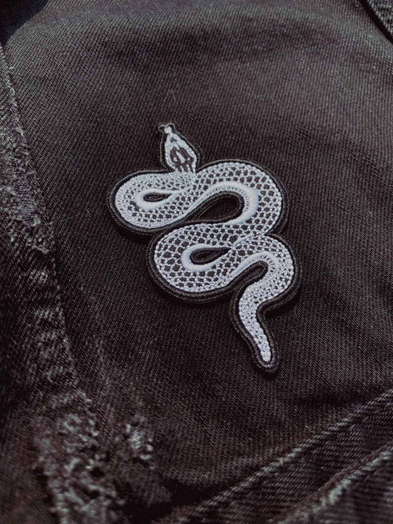 May include: A white snake embroidered patch on a black denim background. The snake is curled up with its head raised.
