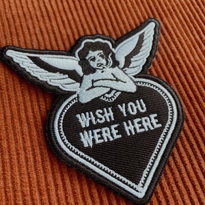 Puede incluir: Un parche negro en forma de corazón con costuras blancas y el texto "Wish You Were Here" en blanco. Un ángel azul con cabello negro está bordado sobre el corazón.