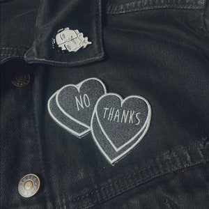 May include: Black denim jacket with two white heart-shaped patches that say "NO" and "THANKS". A silver pin with a black and white illustration of two faces is on the collar.