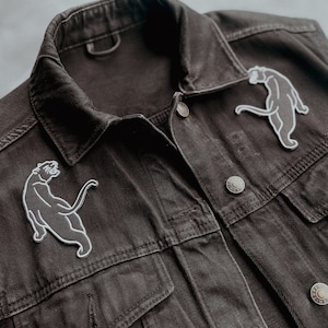 Op de afbeelding: Een zwart denim jack met twee witte geborduurde panter patches op de voorkant.