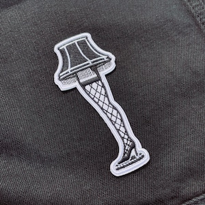 May include: A white embroidered patch of a lampshade with a leg wearing fishnet stockings and a high heel shoe.