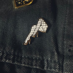 Op de afbeelding: Twee emaille pins op een donkere denim ondergrond. Eén pin is een gouden hart met een huilend gezicht. De andere pin is een gouden en witte pin van een been in visnetkousen en hoge hakken.