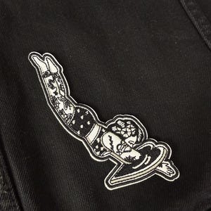 Op de afbeelding: Zwart-wit geborduurde patch met een vrouw die duikt met een surfboard. De patch heeft gedetailleerde tatoeages op de benen en armen. De patch is geplaatst op een zwarte denim achtergrond.