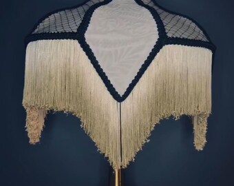 May include: A vintage-style lampshade with a cream-colored fabric and a black net overlay. The lampshade features a diamond shape with a fringe trim. The lamp base is gold-colored.