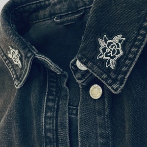May include: A close-up shot of a black denim jacket featuring two embroidered rose patches on the collar. The roses are white with black outlines and green leaves. A silver button is visible.