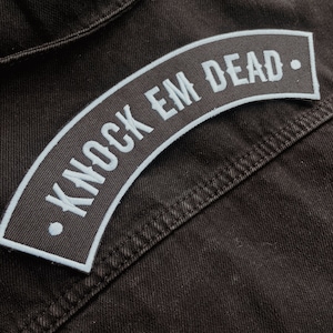 Peut inclure: Une veste en jean noire avec un patch brodé blanc qui dit "Knock 'em Dead."