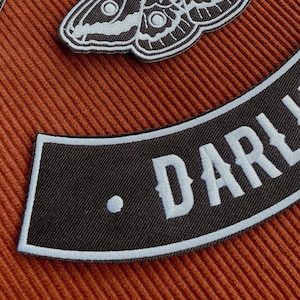 May include: Black fabric patch with white stitching, shaped like a curved banner. The text "DARLING." is embroidered in white on the patch.