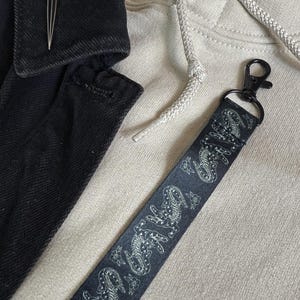 Puede incluir: Lanyard negro y blanco con un clip plateado. El lanyard tiene un diseño repetitivo de criaturas estilizadas.