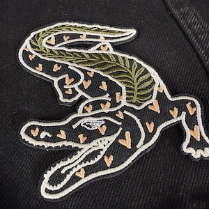 May include: Embroidered alligator patch with a black body, white outline, and peach heart-shaped spots. The alligator has a green leaf design on its back and is set against a black background.