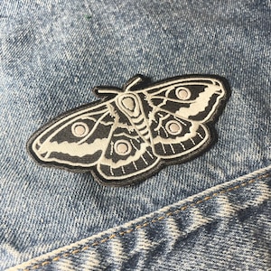 May include: Black and white embroidered moth patch with white eye spots on a blue denim background.