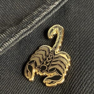 Może przedstawiać: Złota metalowa spinka z czarnym wzorem skorpiona. Skorpion ma szczegółowe ciało i ogon z szeregiem czarnych serc na grzbiecie.