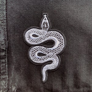 May include: A white embroidered snake patch on a black denim background.