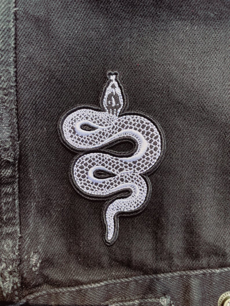 May include: A white embroidered snake patch on a black denim background.
