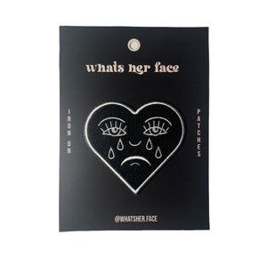 Puede incluir: Parche termoadhesivo blanco y negro con forma de coraz&oacute;n y una cara llorando con pesta&ntilde;as. El texto "whats her face" est&aacute; en la parte superior del parche y "@whatsher.face" en la parte inferior.