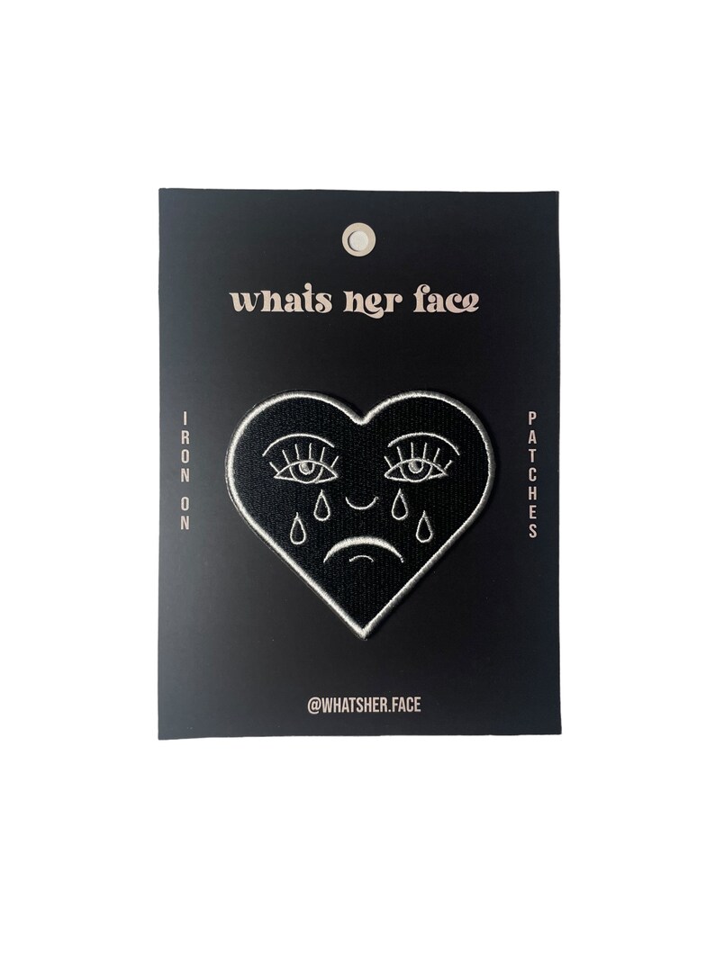Puede incluir: Parche termoadhesivo blanco y negro con forma de coraz&oacute;n y una cara llorando con pesta&ntilde;as. El texto "whats her face" est&aacute; en la parte superior del parche y "@whatsher.face" en la parte inferior.