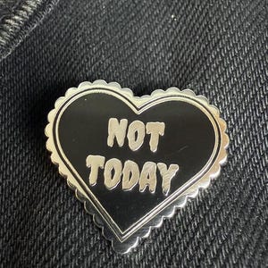 Puede incluir: Pin de esmalte negro en forma de corazón con borde plateado y el texto "NOT TODAY" en blanco.