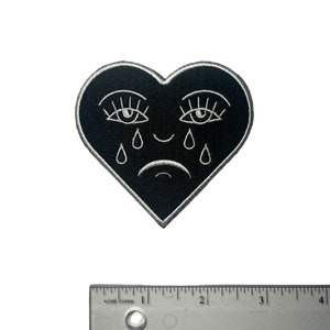 Puede incluir: Parche de fieltro negro en forma de coraz&oacute;n con ojos, pesta&ntilde;as y l&aacute;grimas bordadas en blanco. El coraz&oacute;n tiene una peque&ntilde;a sonrisa.