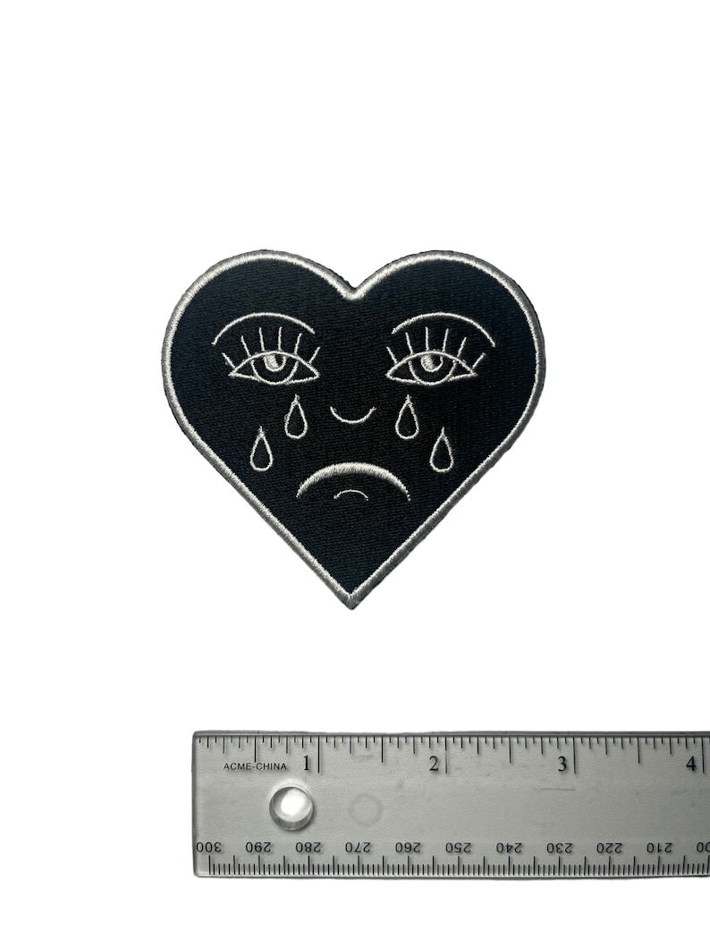 Puede incluir: Parche de fieltro negro en forma de coraz&oacute;n con ojos, pesta&ntilde;as y l&aacute;grimas bordadas en blanco. El coraz&oacute;n tiene una peque&ntilde;a sonrisa.