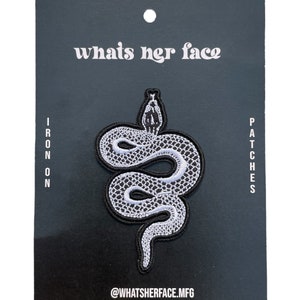 May include: Iron-on patch featuring a white snake with black outlines on a black background. The patch is packaged on a black card with the text "whats her face" and "@whatsherface.mfg".