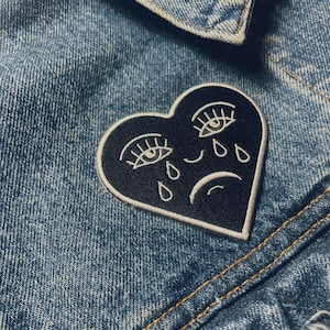 Puede incluir: Un parche negro en forma de coraz&oacute;n con ojos, pesta&ntilde;as y l&aacute;grimas bordadas en blanco. El coraz&oacute;n tiene un contorno blanco y una l&iacute;nea curva blanca que representa una mueca.