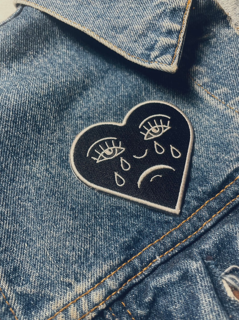 Puede incluir: Un parche negro en forma de coraz&oacute;n con ojos, pesta&ntilde;as y l&aacute;grimas bordadas en blanco. El coraz&oacute;n tiene un contorno blanco y una l&iacute;nea curva blanca que representa una mueca.