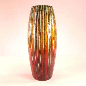 May include: A tall, cylindrical ceramic vase featuring a vertical striped pattern. The design transitions from a deep red base to a warm yellow and orange midsection, accented by thin, light green vertical lines and dark brown detailing.