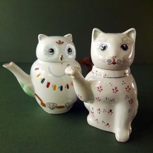 May include: Two ceramic teapots, one shaped like an owl and the other like a cat. The owl teapot is white with colorful accents and a detailed face. The cat teapot is also white, with a floral pattern and a raised paw. Both have lids.