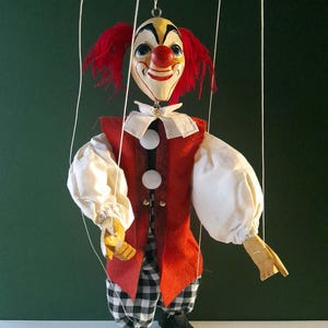 May include: A vintage clown puppet with a painted face, red hair, and a red and white outfit. The puppet has a red vest, white sleeves, and black and white checkered pants. It is suspended by strings.