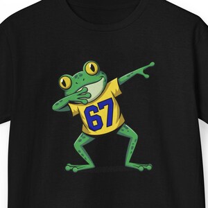 May include: Black t-shirt featuring a green cartoon frog dabbing. The frog wears a yellow shirt with the number 67 in blue. The frog has large, yellow-rimmed eyes and is in a dabbing pose.