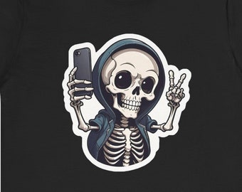 Puede incluir: Una camiseta negra con un esqueleto de dibujos animados que lleva una sudadera azul y se hace un selfie con un teléfono inteligente negro. El esqueleto está haciendo un signo de paz con una mano.