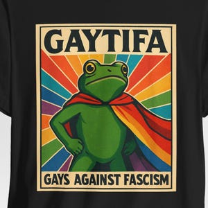 May include: Black t-shirt with a green frog superhero wearing a rainbow cape, with the text "GAYTIFA GAYS AGAINST FASCISM." The frog is set against a rainbow striped background. The design is printed on a black t-shirt.