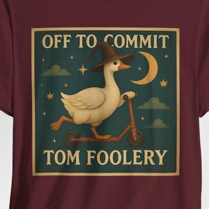 May include: Burgundy t-shirt featuring a whimsical graphic. The graphic depicts a goose wearing a witch hat, riding a scooter under a crescent moon and stars, with the text "OFF TO COMMIT TOM FOOLERY".